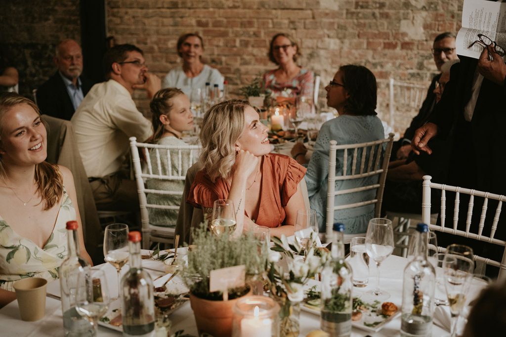 Eine Frau sitzt an einer Hochzeitstafel, im Hintergrund weitere Gäste
